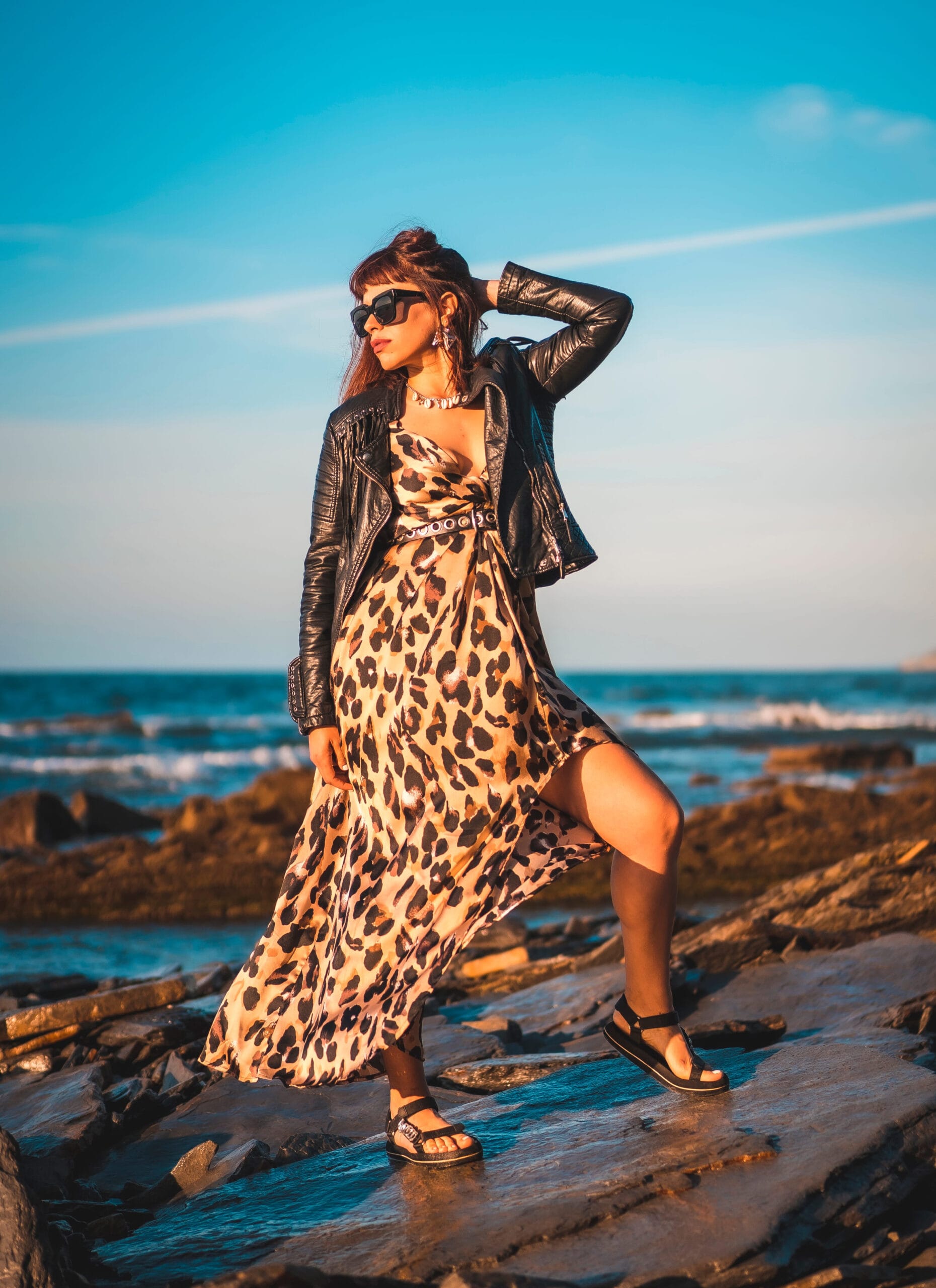 Summer street style on a beach next to rocks of a young brunette caucasian woman in a leopard dress by the sea.
